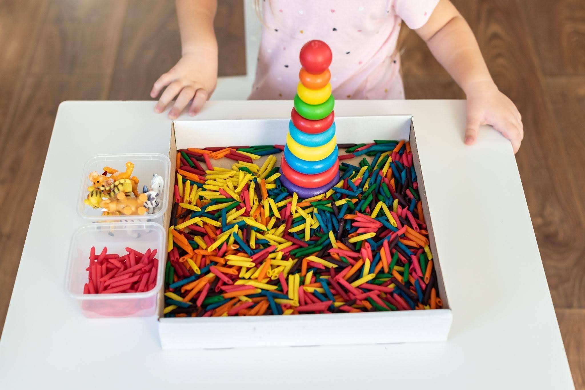 Child stacks a colorful rainbow ring tower inside a box filled with multicolored pasta pieces as a pretend play activity.