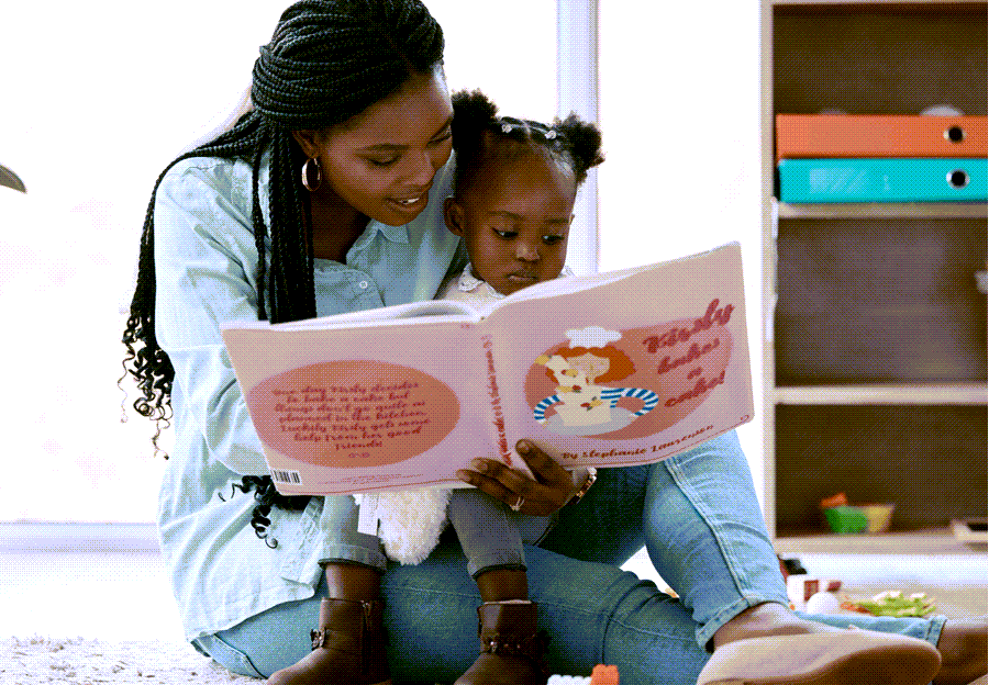 A woman with braided hair reads a pink children's book to a young child while sitting on the floor.